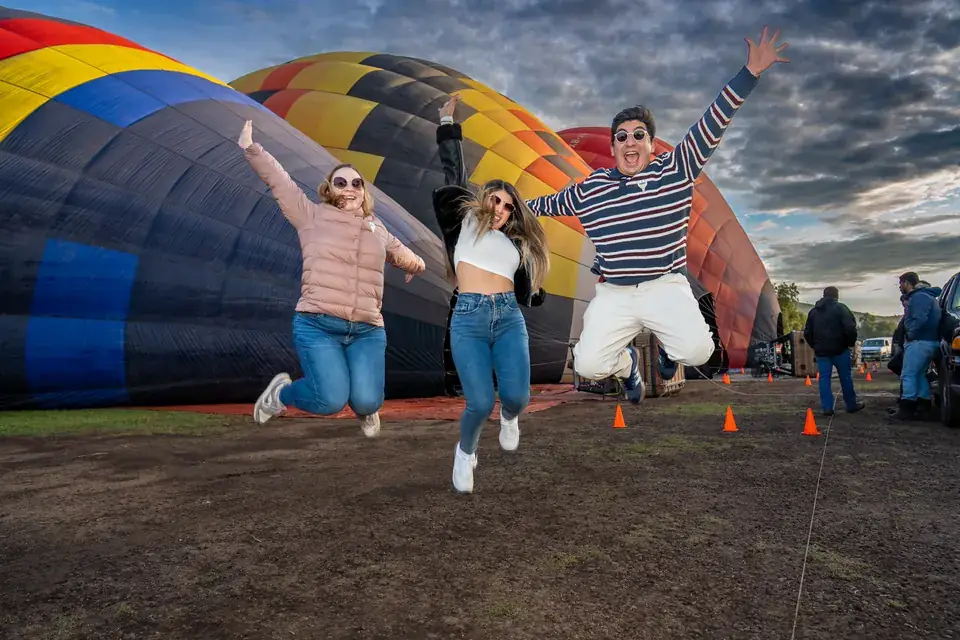 Three friends jump for joy in front of hot air balloons inflating at sunrise in Teotihuacan