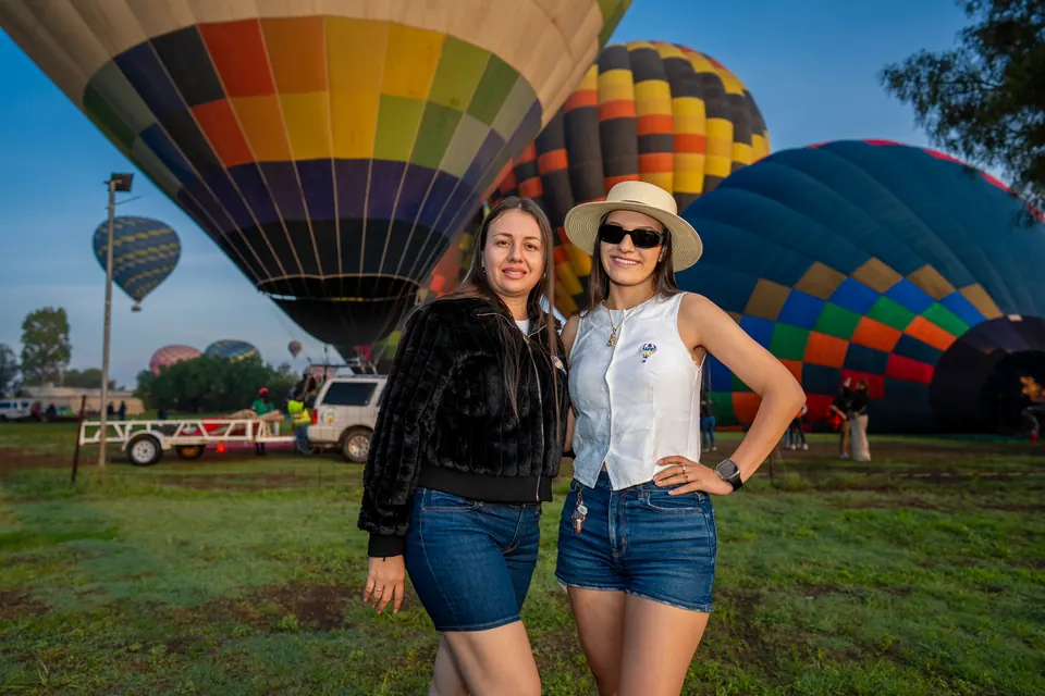 Two smiling friends in front of inflated hot air balloons on the grass at sunrise