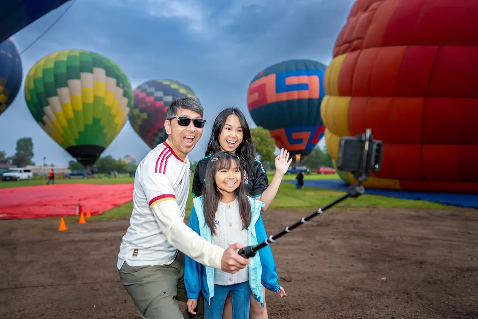 Smiling family taking a selfie with several inflated hot air balloons behind them at the Teotihuacan launch field