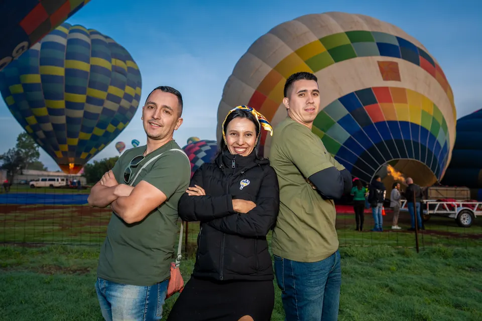 Tres amigos posando sobre el pasto frente a dos globos aerostáticos inflados al amanecer