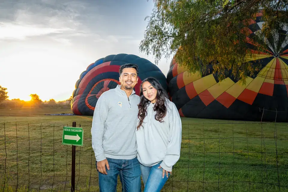 Smiling couple side by side in the green field as two balloons inflate behind them at sunrise