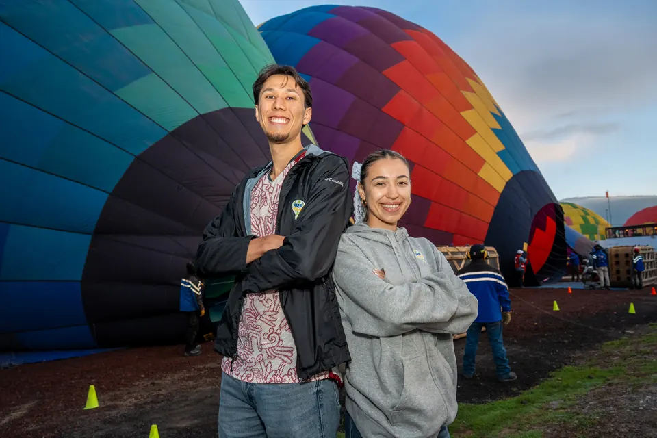 Smiling couple standing back to back with folded arms as a huge balloon inflates behind them